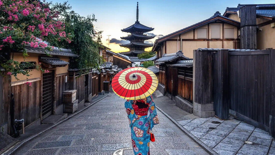 Femme en kimono avec un parapluie traditionnel marchant dans une rue de Kyoto, avec une pagode en arrière-plan au coucher du soleil.