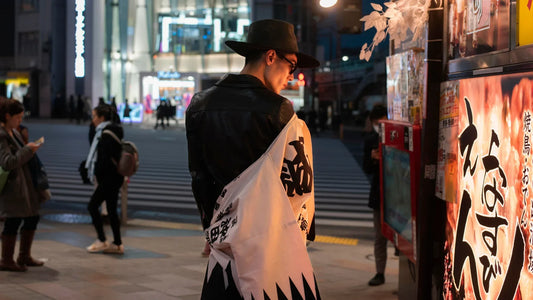 Homme en tenue streetwear japonais avec un sweat manga et un chapeau noir, photographié de nuit dans une rue animée de Tokyo.