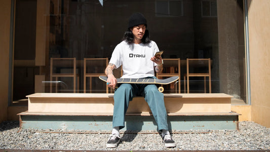 Homme portant un t-shirt streetwear japonais blanc avec l'inscription Otaku, assis avec un skateboard en main devant une vitrine urbaine.