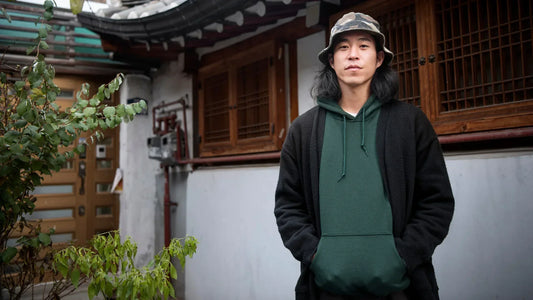 Homme portant un sweat à capuche vert et un cardigan noir, posant devant une maison traditionnelle japonaise, représentant la fusion entre culture urbaine et héritage nippon.